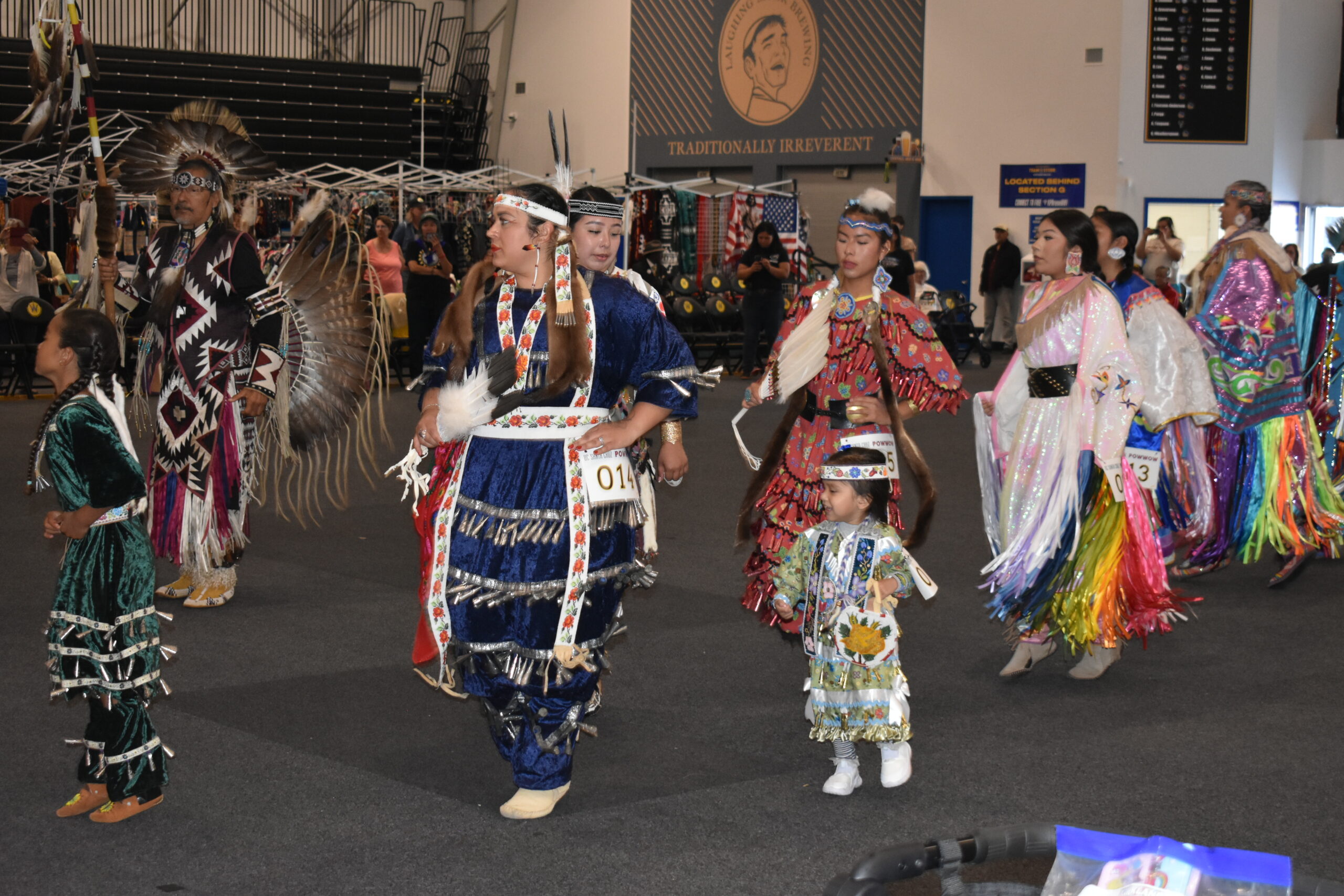 A multigenerational group of Indigenous dancers in colorful, traditional regalia participate in a dance at the UCSC Powwow.